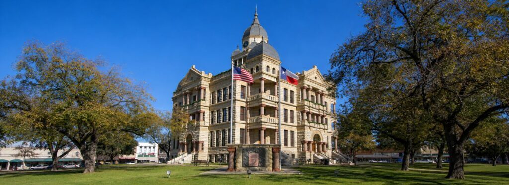 Denton County Courthouse -Capitol-style stone building with a dome, flags flying, set in a sunny park with trees on both sides.
